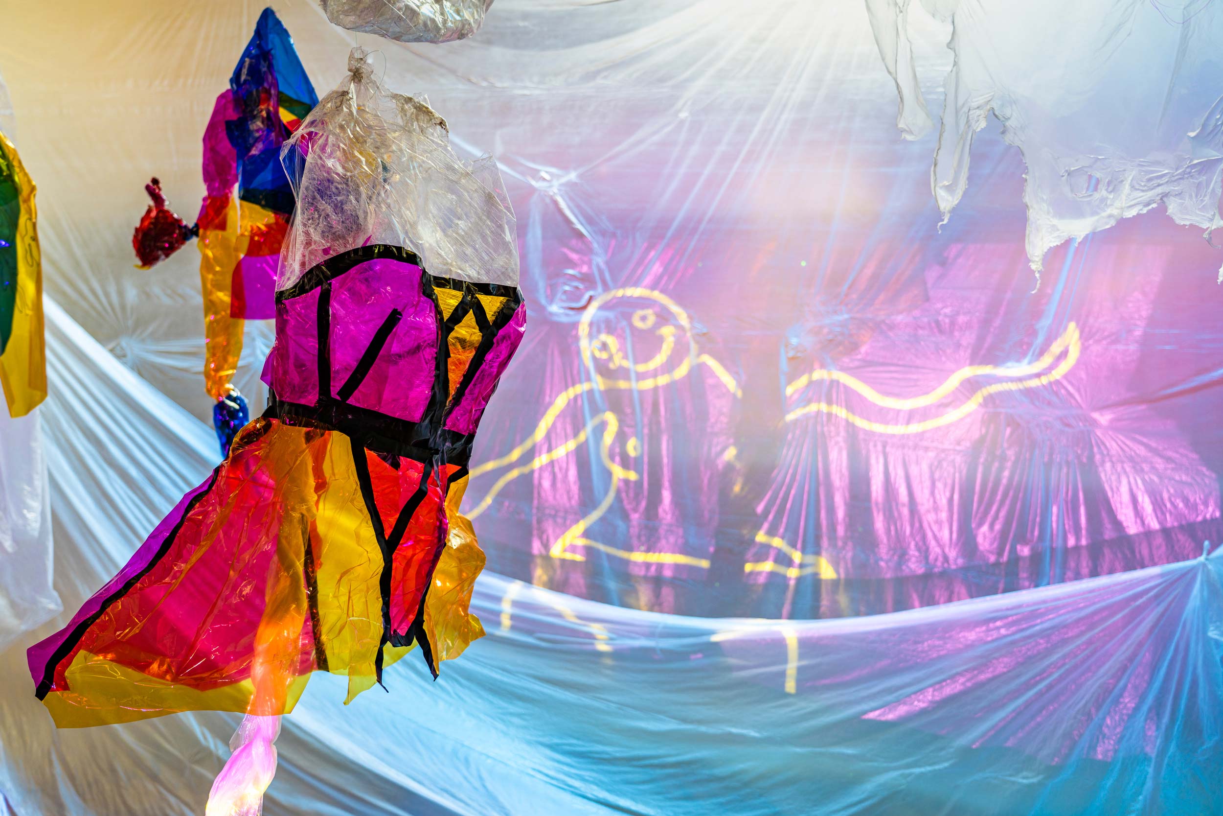 Installation featuring pink and yellow transparent plastic corset/dree hanging. An animation of a female character drawn in yellow is projected onto the purple and blue plastic background.