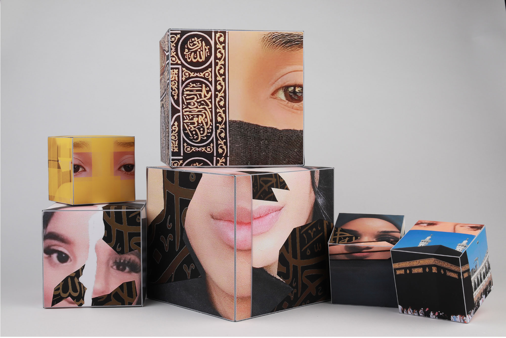 A series a cubes with photographs printed on them are stacked against a white background. The cubes show a young woman eyes and mouth, alongside architectures from the Middle East. The female characters represented on the cube appear to wear a head scarf.
