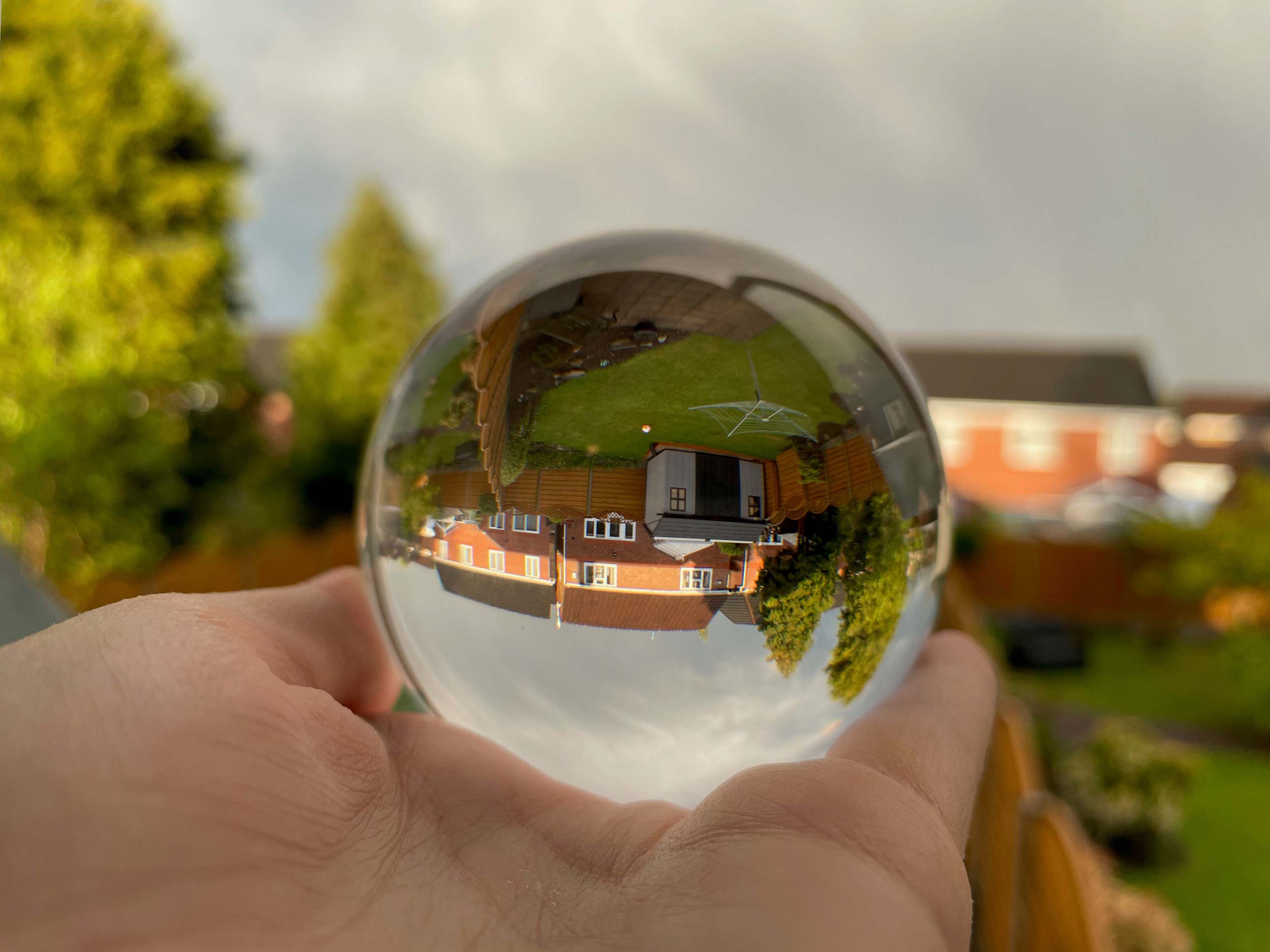 A colour photograph showing A hand is holding a glass sphere. Inside the sphere a house is reflected. It is upside down.