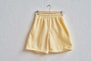 Photograph of yellow shorts made from soft towelling fabric hanging on a wooden coat hanger on a white wall.