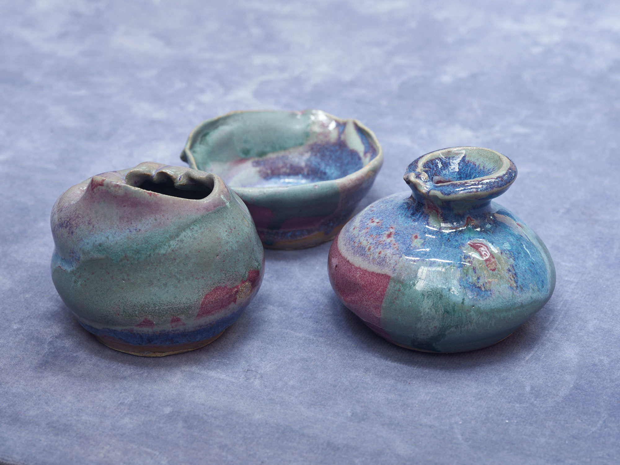 a photograph of three ceramic pots on a light blue fabric. the ceramics are odd shaped vases and are glazed in green , blue and red.