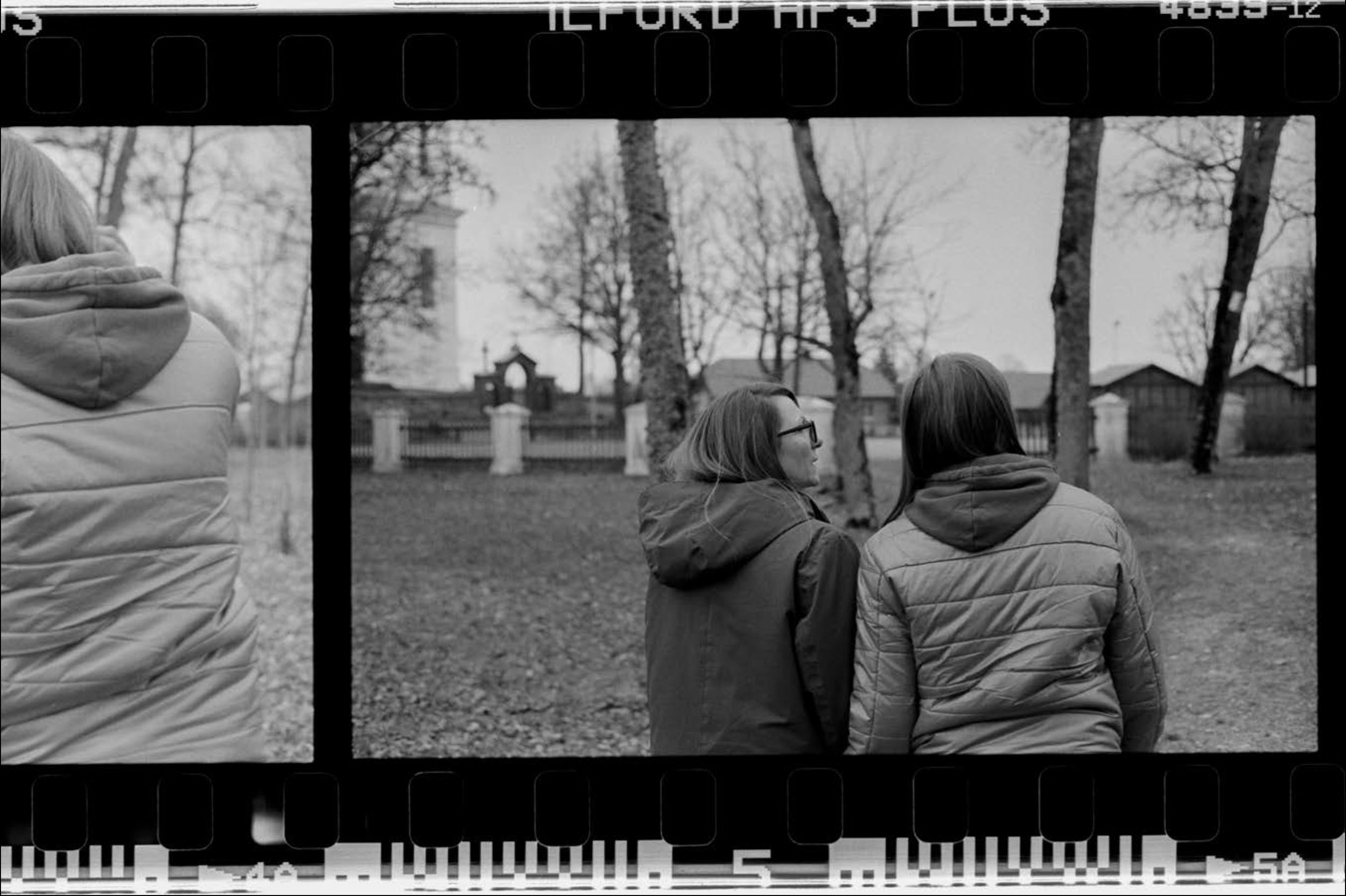 A black and white photograph shows the backs of two women as they walk towards a series of trees and small buildings in the distance. The photograph is a scan from the analogue film and details of the film can be seen at the top and bottom of the image, including the words Ilford HP5 Plus at the top and a series of light grey graphic shapes at the bottom.