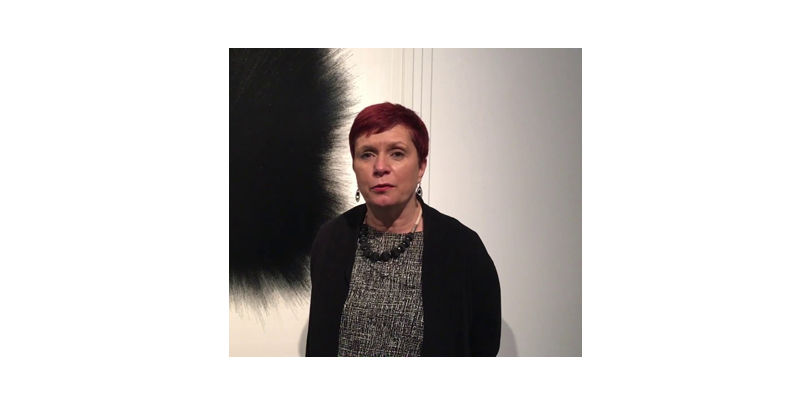 Deborah Robinson standing in front of a painting. Deborah is a white woman with short hair. She is wearing a grey top and black cardigan/jacket.