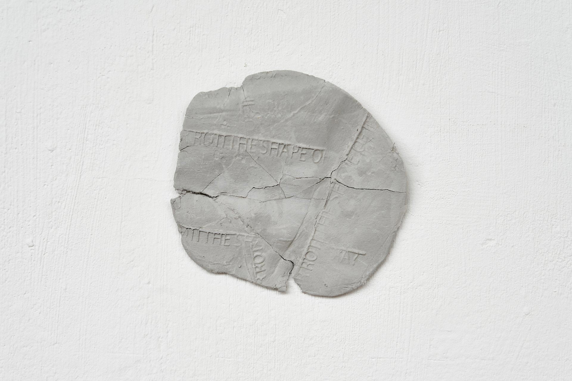 grey circle shaped clay disk on a white wall. The grey circle has the words 'Rotti the shape of' printed onto it.