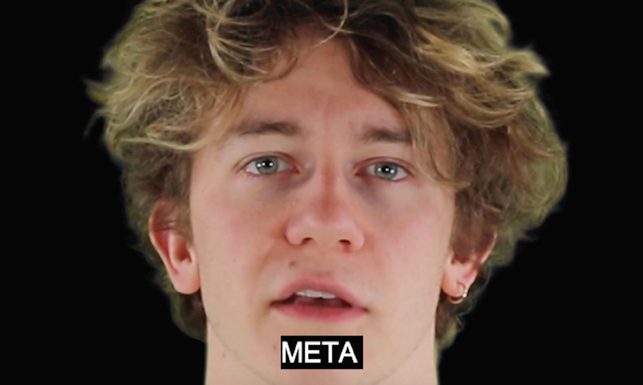 A film still showing a black background with a young males face. He is white and has medium length brown hair. The word 'Meta' is written at the bottom of the screen